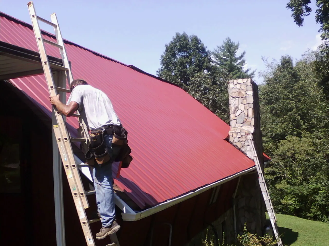 Roofer on a ladder beside a red metal roof and stone chimney in Upperville
